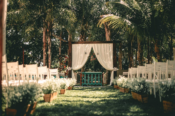 Beautiful lush palm trees overhang white chair separated by a grass aisle. A wooden frame with drawn back curtains is at the top of the aisle.